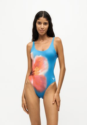 Femme aux longs cheveux foncés portant un maillot de bain une pièce bleu avec un grand motif de fleurs roses et oranges à l'avant.