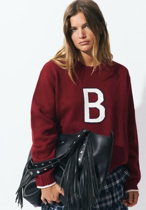 Suéter burdeos de punto con una gran "B" blanca en la parte frontal, combinado con un bolso de cuero negro con flecos y detalles de tachuelas.