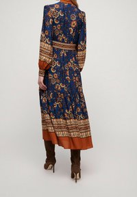 Långärmad klänning i blått med blommönster, har en åtsittande midja och bruna detaljer. Funktioner inkluderar en plisserad kjol och kontrasterande fåll.