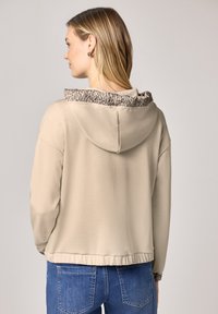 Kvinde med lys hud og blondt hår, iført beige hættetrøje med leopardmønstret foer i hætten, kombineret med blå denim-jeans, set fra venstre side og bagfra.