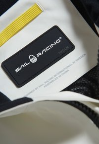 Svart och vit inredning med ett gult accentband; har en svart etikett med "SAIL RACING"-logotyp, storlek "MEDIUM" och skötselinformation.