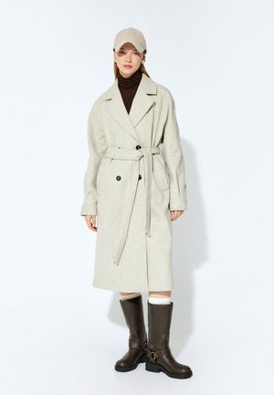 Pitkä beige takki, jossa on vyötärölle belted, mustat kaksinkertaiset napit ja ylisuurikokoinen kaulus, yhdistetty tummanruskeisiin polvipitkiin saappaisiin ja beigeen lippalakkiin.