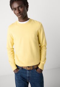 Sárga környakú pulóver, pamutból készült, slim fit fazonnal és egy kis logóval. Kék farmernadrággal és barna övvel viselve.