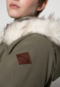 Mujer con chaqueta verde oliva con cuello de piel sintética blanca y parche marrón de la marca Hollister en la parte superior de la manga.