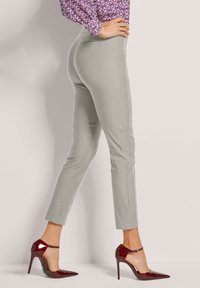 Frau in hellgrauen, schmal geschnittenen Knöchellhosen und bordeauxfarbenen, lackledernen High Heels, seitlich stehend mit den Händen in der Hüfte.