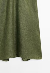 Ourlet en tissu de lin vert olive avec une texture visible, suspendu sur un fond clair.