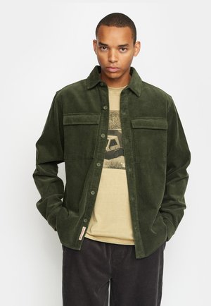 Dark green corduroy shirt jacket with two chest pockets, button front, classic collar, and a beige graphic t-shirt underneath.