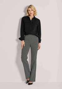 Vrouw staat tegen de muur gekleed in een zwart overhemd met knopen, gestreepte zwart-witte broek en zwarte puntige hakken.