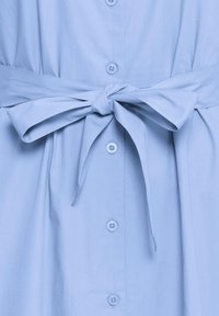 Lyseblå skjortebluse med stofbælte bundet i en sløjfe i taljen, placeret midt for på forsiden.
