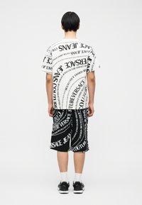 Άτομο που στέκεται με την πλάτη, φορώντας μαύρο και λευκό t-shirt και σορτς Versace Jeans Couture με μοτίβο κειμένου λογότυπου σε όλη την επιφάνεια.