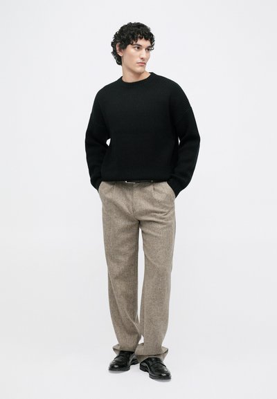 Zwarte gebreide trui met ribbeltextuur, gecombineerd met beige geruite broek en zwarte schoenen. Model staat met de handen in de zakken.