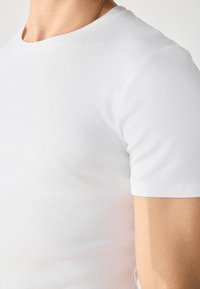 Bílý bavlněný tričko s přiléhavým střihem, žebrovanou texturou a krátkými rukávy. Obsahuje kulatý výstřih a bezšvová ramena.