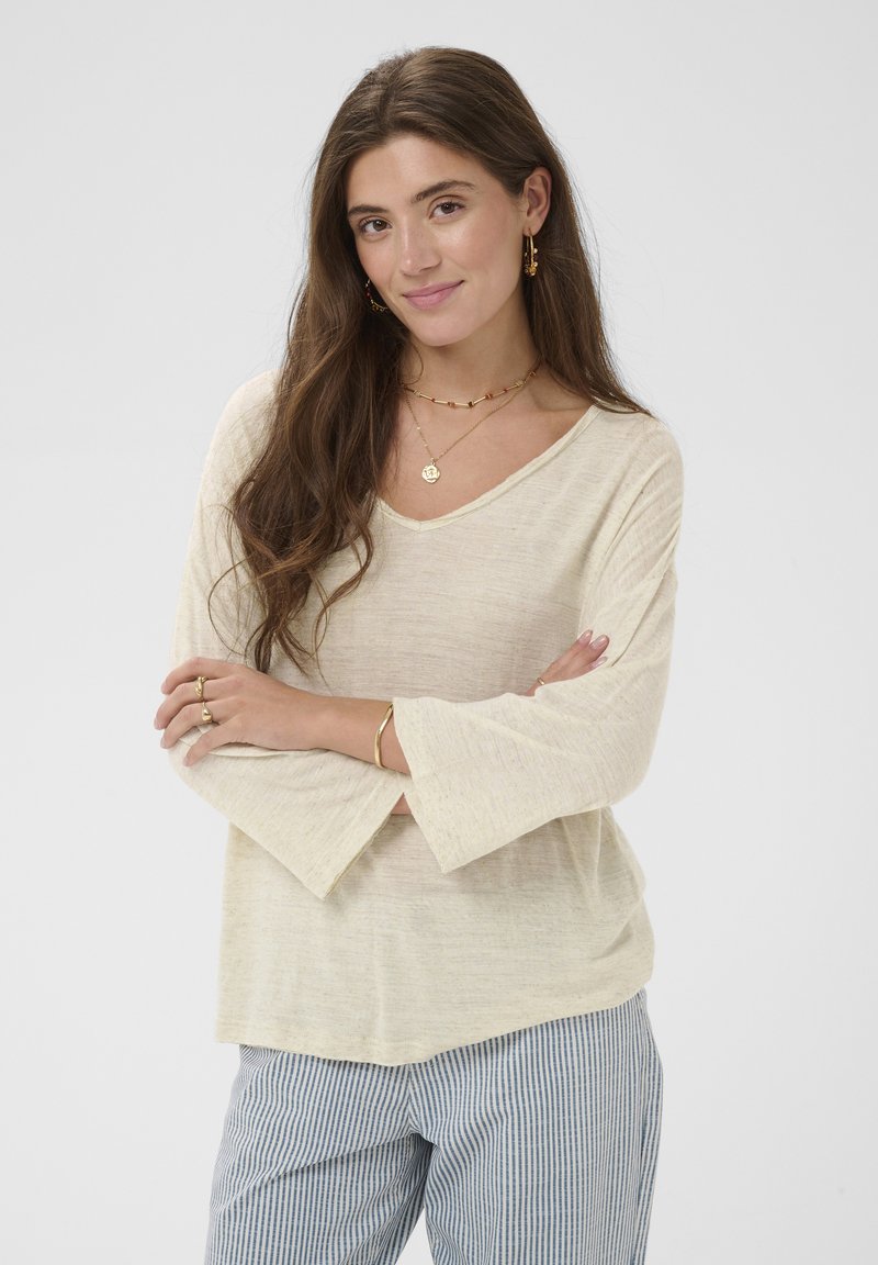 Femme aux longs cheveux bruns portant un haut ample à manches longues beige et un pantalon rayé bleu, les bras croisés, souriant doucement devant un fond uni.