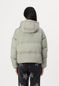 Femme aux longs cheveux bruns portant une doudoune grise claire avec capuche et un jean noir orné de patchs étoiles argentés, vue de dos.