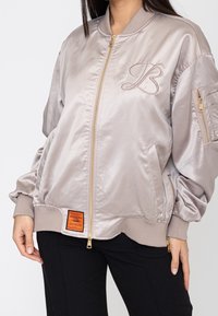 Femme portant une veste bomber beige brillante avec fermeture éclair frontale, logo brodé sur la poitrine, patch orange près de l’ourlet et poche zippée sur la manche.
