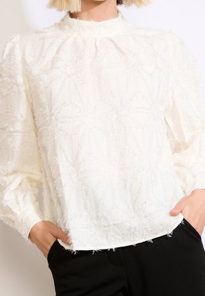 Mujer con blusa blanca texturizada de manga larga y escote alto, combinada con pantalones negros, manos en los bolsillos.