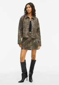 Veste raccourcie et mini jupe en imprimé serpent brun texturé. Modèle portant des bottes noires à talons aiguilles jusqu'aux genoux. Tissu lisse avec des boutons métalliques.