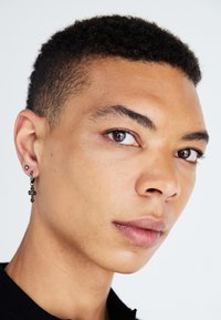 Sort korsformet ørering med en glitrende accent, båret i et piercet øre. Nærbillede af en persons ansigt med kort krøllet hår og neutral hudtone.