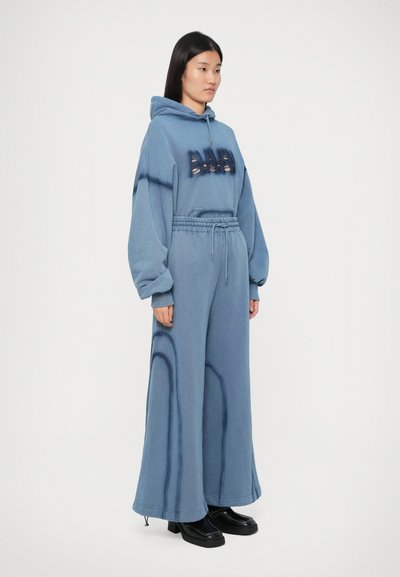 Žena u plavoj oversized dukserici i odgovarajućim širokim hlačama, s crnim platformskim cipelama, stoji ispred bijele pozadine.