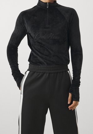 Top in velluto nero a maniche lunghe con colletto a zip, caratterizzato da un logo ricamato. Abbinato a pantaloni jogger neri con strisce laterali bianche.