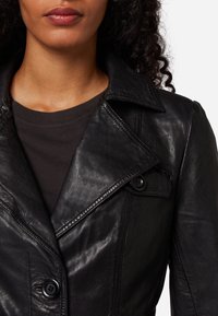 Kvinna som bär en svart läderjacka med breda slag över en mörk tröja med rund hals, där hals och lockiga hårslingor syns.