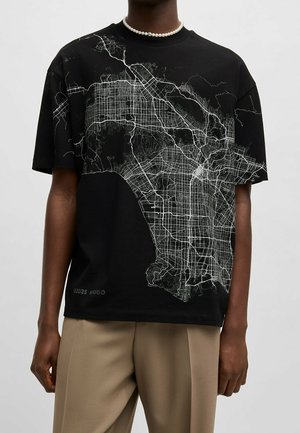 Persoon draagt een zwart t-shirt met een gedetailleerd wit kaartontwerp, beige broek en een parelketting tegen een effen achtergrond.