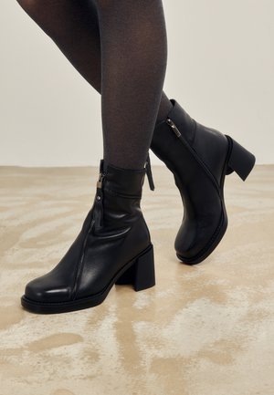 Beine in dunklen Strumpfhosen, die schwarze Leder-Stiefeletten mit seitlichen Reißverschlüssen und Blockabsätzen auf einem hell gemusterten Boden tragen.
