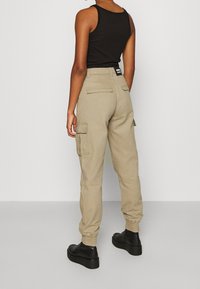 Béžové cargo kalhoty s zadními a bočními kapsami, elastickými manžetami a uvolněným střihem, kombinované s černým tílkem a černými botami.