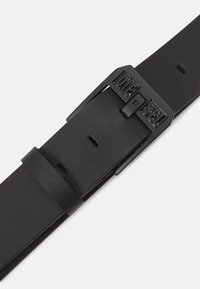 Curea din piele neagră cu o cataramă metalică mată, inscripționată cu textul "DIESEL", textură netedă și mai multe găuri de ajustare pe curea.
