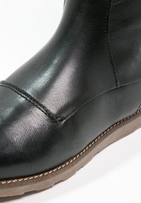 Bota de cuero negro hasta el tobillo con costuras visibles y una suela de goma marrón, mostrada en primer plano sobre un fondo blanco.