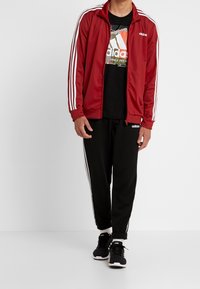 Chaqueta de chándal roja con cremallera y rayas blancas, camiseta negra con logo, pantalones de chándal negros y zapatillas negras con detalles blancos.