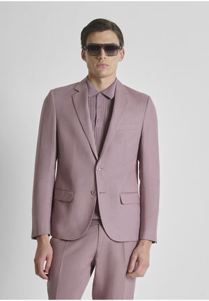 Man draagt een mauve pak met bijpassend gesloten overhemd en grote donkere zonnebril, staande tegen een effen lichte achtergrond.