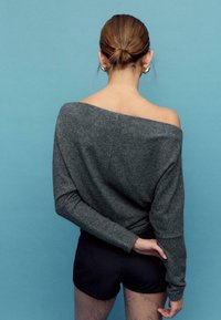 Siv pulover z odkritimi rameni iz mehke tkanine, z dolgimi rokavi in sproščenim krojem, kombiniran s črnimi kratkimi hlačami in vzorčastimi najlonkami.