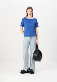 Blå kortermet topp med trykket tekst, kombinert med høydejusterte lyse jeans med naglede detaljer. Modellen holder en svart veske.