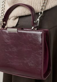 Majhna bordo usnjena torbica z teksturirano površino, zgornjim ročajem in srebrno verižico. Ponaša se z rektangularno kovinsko zaponko.