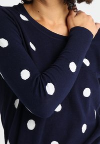 Granatowa, dziana bluzka z długim rękawem w białe kropki, okrągły dekolt i prążkowane mankiety. Miękka faktura, casualowy fason.