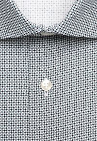 Gros plan sur un col de chemise et une patte avec un petit motif géométrique en forme de losange noir, blanc et bleu clair, avec un bouton blanc.