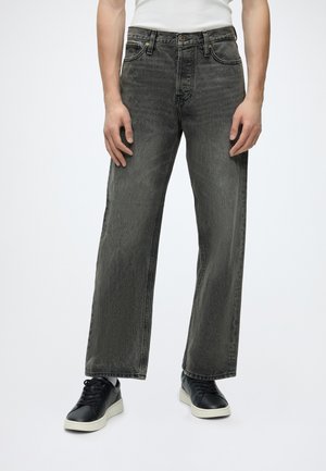Personne portant un jean noir délavé droit, des baskets noires avec des semelles blanches et un haut blanc rentré, debout devant un fond blanc.
