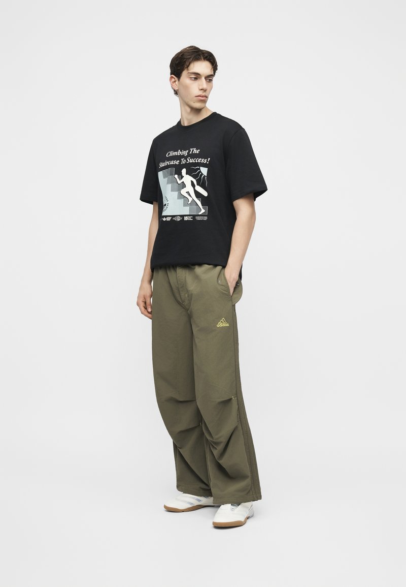 Junger Mann in schwarzem Grafik-T-Shirt mit der Aufschrift „Climbing The Staircase To Success!“ und olivgrünen, weit geschnittenen Adidas-Hosen mit weißen Sneakers.