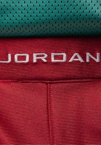 Gros plan d'un tissu rouge avec une broderie blanche "JORDAN" et un tissu en mesh bleu sarcelle plié avec de petits trous de ventilation au-dessus.
