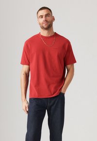 Rød bomulds T-shirt med korte ærmer, rund hals og diskret logo-detaljering. Båret med mørkeblå jeans, der har en glat tekstur.