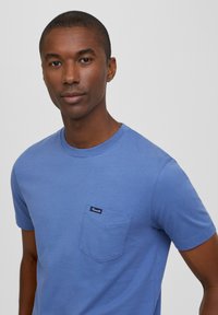 T-shirt in cotone blu con scollatura rotonda e maniche corte. Presenta una tasca sul lato sinistro del petto con un'etichetta nera. Texture liscia.