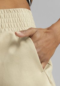 Beige sportovní kalhoty s nařaseným pasem a bočními kapsami, s hladkým, elastickým povrchem a detailním záběrem ruky uvnitř kapsy.