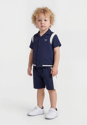 Κατσαρός νήπιος που φοράει πουκάμισο με κοντά μανίκια σε ναυτικό μπλε και σορτς που ταιριάζει με λευκές πλευρικές ρίγες και λευκά αθλητικά παπούτσια, όρθιος σε λευκό φόντο.