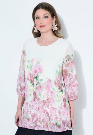 Femme portant un chemisier blanc plissé avec un imprimé floral rose et des manches trois-quarts, associé à de grandes boucles d'oreilles en forme de fleur rose.