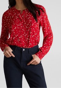 Femme portant un blouse rouge à manches longues avec un imprimé floral blanc, rentrée dans un pantalon sombre marine taille haute, les mains dans les poches.