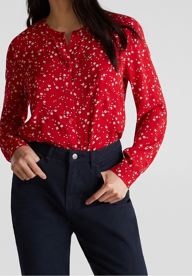 Femme portant un blouse rouge à manches longues avec un imprimé floral blanc, rentrée dans un pantalon sombre marine taille haute, les mains dans les poches.