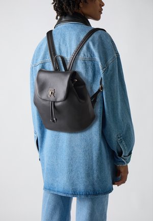 Schwarzer Leder-Rucksack mit einer runden Klappe, Zugbandverschluss und geprägtem Logo. Wird über einer Jeansjacke getragen. Glatte Textur und lässiges Design.
