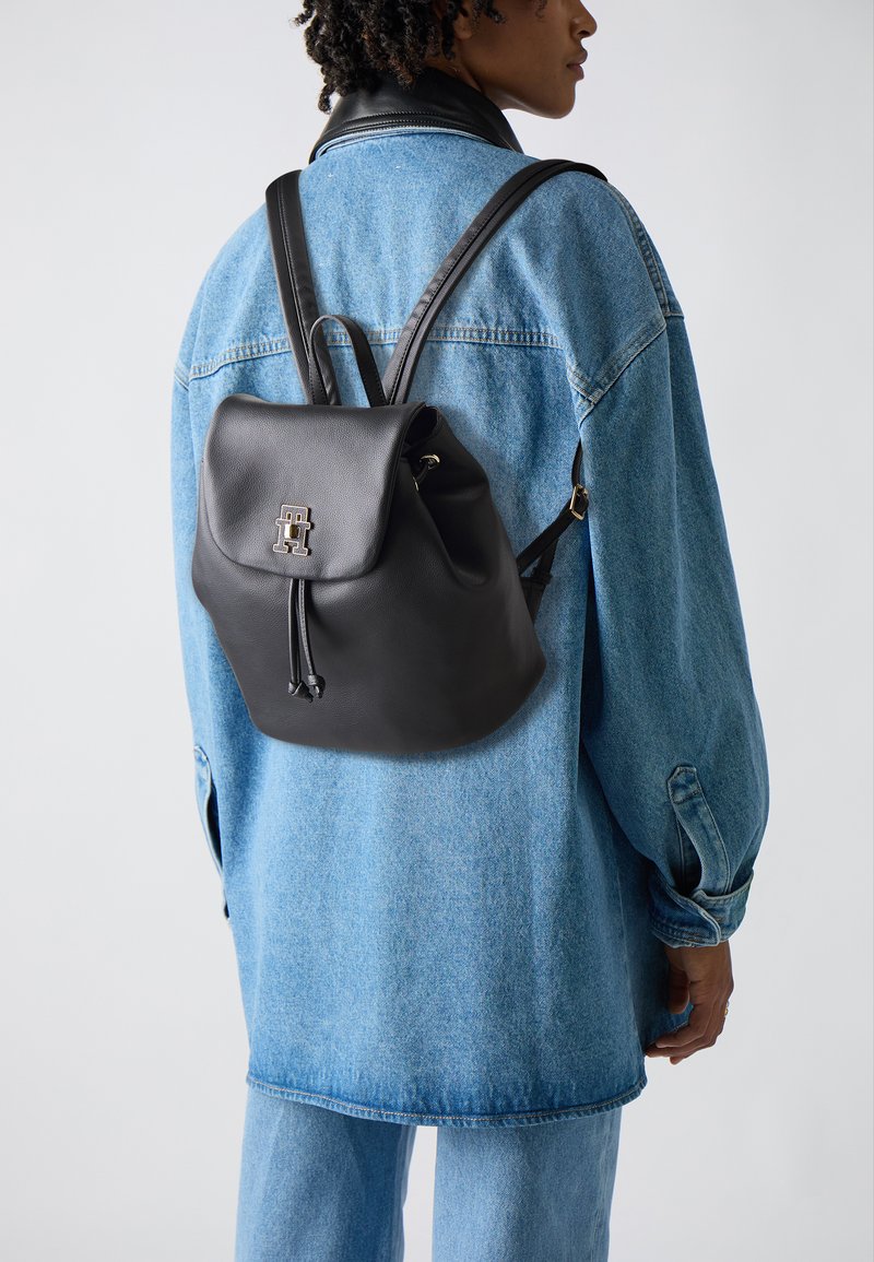 Schwarzer Leder-Rucksack mit einer runden Klappe, Zugbandverschluss und geprägtem Logo. Wird über einer Jeansjacke getragen. Glatte Textur und lässiges Design.