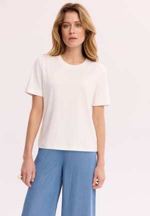 Kobieta o blond włosach do ramion, ubrana w jednolity biały t-shirt z krótkim rękawem i jasnoniebieskie spodnie z szerokimi nogawkami, stojąca na neutralnym tle.